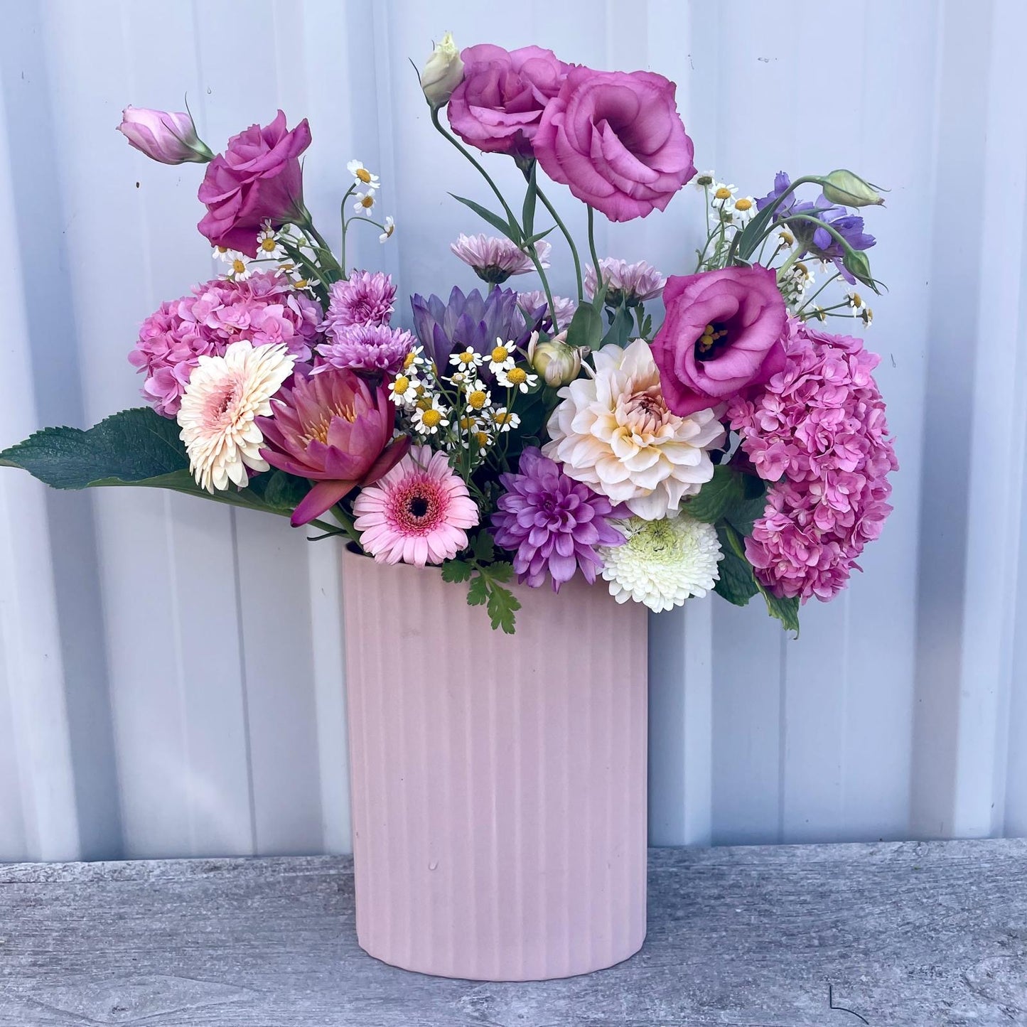 Vibrant seasonal flowers burst with color in a soft pink tin vase, creating a cheerful garden arrangement for any space.