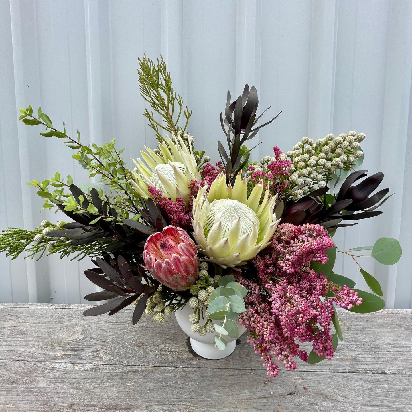 Vibrant deluxe native arrangement featuring three bold king proteas, wildflowers, and lush foliage in a tall glass vase with elegant ribbons.