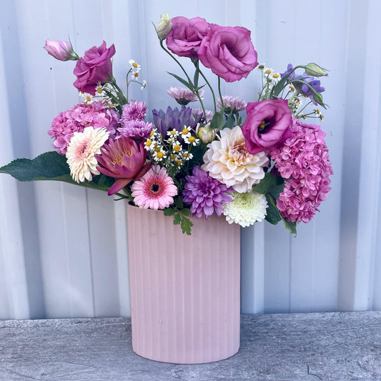 Vibrant seasonal flowers burst with color in a soft pink tin vase, creating a cheerful garden arrangement for any space.