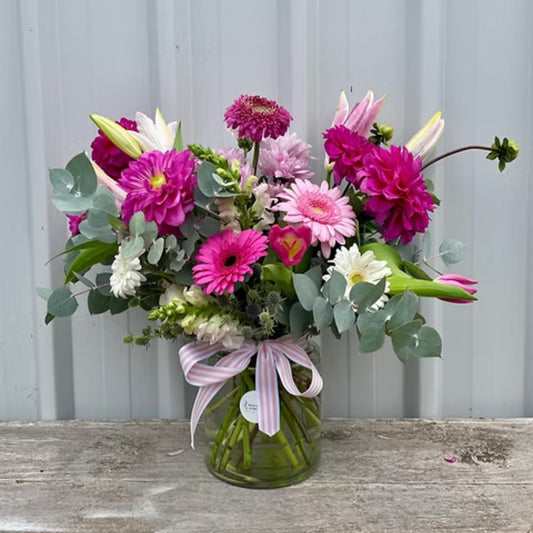 Vibrant medium seasonal flower arrangement in a 20cm glass vase, featuring gerberas, lilies, and colorful blooms with a ribbon accent.