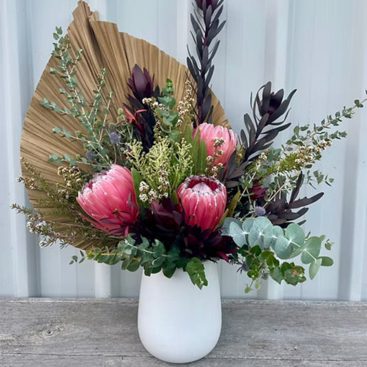 Vibrant locally grown flowers, including bold king proteas and wildflowers, arranged with dried fan palm in a tall ceramic vase.