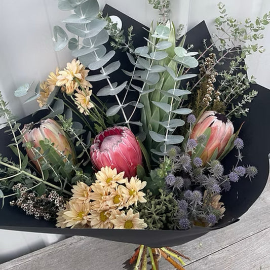 Vibrant large bouquet of native flowers with a striking king protea, colorful pincushions, and wildflowers, wrapped in rustic brown paper with a matching ribbon.