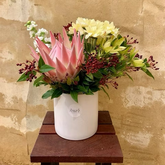 Vibrant small native arrangement with a striking king protea, colorful local flowers, and lush foliage in a chic white ceramic pot.