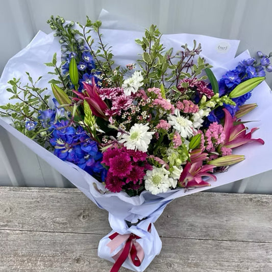 Vibrant Deluxe Seasonal Bouquet with lilies, snapdragons, and gerberas, wrapped in brown paper with elegant ribbons.