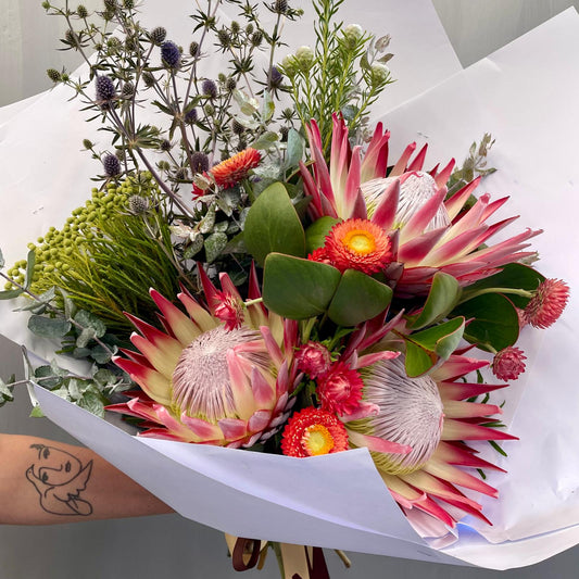 Stunning Deluxe Native Bouquet with vibrant king proteas, wildflowers, and lush foliage, wrapped in brown paper with matching ribbons.
