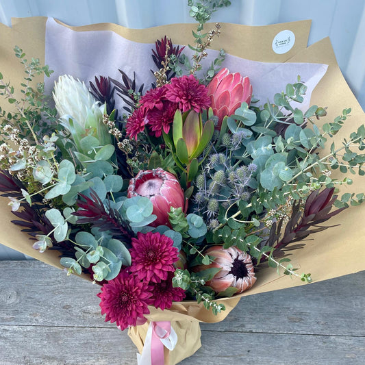 Vibrant bouquet of locally grown native flowers with a striking king protea, colorful wildflowers, and lush foliage, wrapped in rustic brown paper with a matching ribbon.