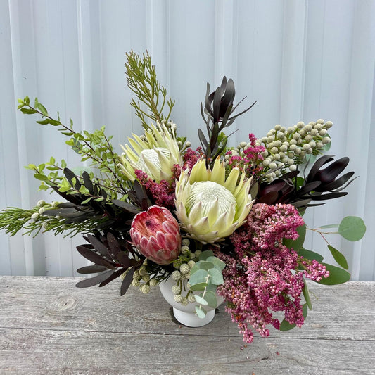 Vibrant deluxe native arrangement featuring three bold king proteas, wildflowers, and lush foliage in a tall glass vase with elegant ribbons.